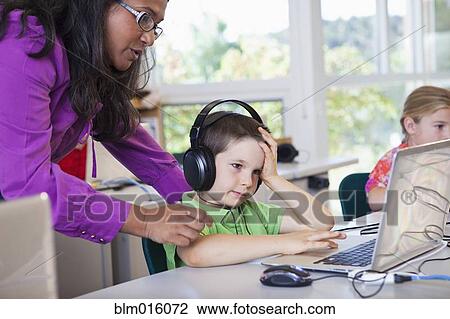 Stock Image - Teacher helping student using laptop in classroom. Fotosearch