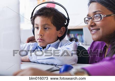 Stock Image - Teacher helping student using laptop in classroom. Fotosearch