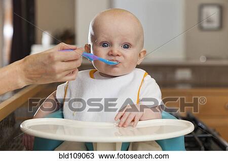 Stock Image - Caucasian mother feeding baby in high chair. Fotosearch
