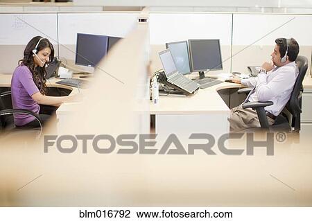 Stock Image - Business people wearing headsets at desks. Fotosearch