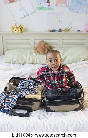 Stock Photography - African American boy sitting in suitcase on bed. Fotosearch