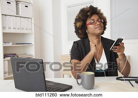 Stock Image - African American businesswoman listening to headphones at desk. Fotosearch