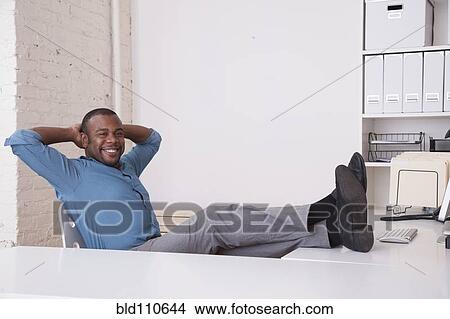 Picture - Black businessman sitting at desk with feet up. Fotosearch