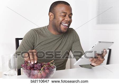 Stock Photo - Black man eating grapes and using digital tablet. Fotosearch