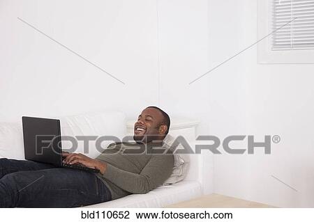 Stock Image - Black man laying on sofa using digital tablet. Fotosearch