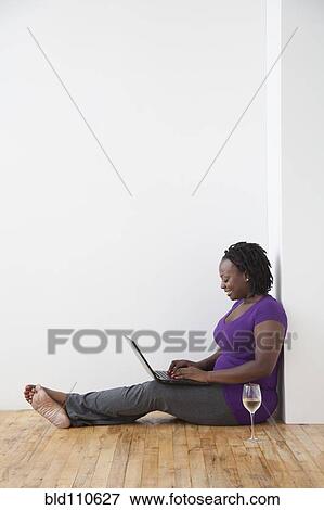 Stock Photo - Black woman drinking wine and using laptop on floor. Fotosearch