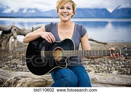 Stock Image - Caucasian woman playing guitar near lake. Fotosearch