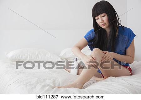 Stock Photo - Chinese woman moisturizing legs on bed. Fotosearch