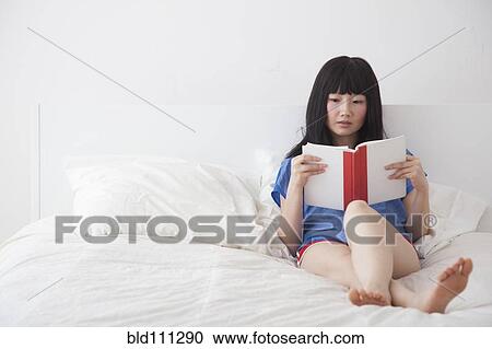 Stock Image - Chinese woman reading book on bed. Fotosearch