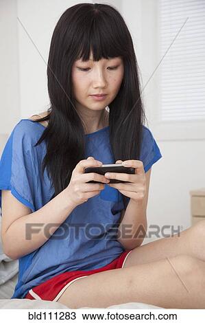 Stock Image - Chinese woman using cell phone on bed. Fotosearch