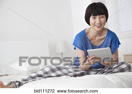 Stock Image - Chinese woman using tablet computer on bed. Fotosearch