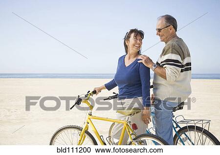 Stock Image - Couple standing with bicycles on beach. Fotosearch