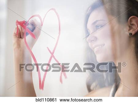 Stock Image - Hispanic girl drawing heart with arrow on window. Fotosearch