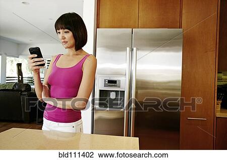 Stock Image - Mixed race woman using cell phone in kitchen. Fotosearch