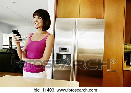 Stock Image - Mixed race woman using cell phone in kitchen. Fotosearch