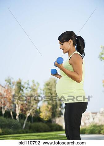 Stock Image - Pregnant Hispanic woman lifting weights outdoors. Fotosearch