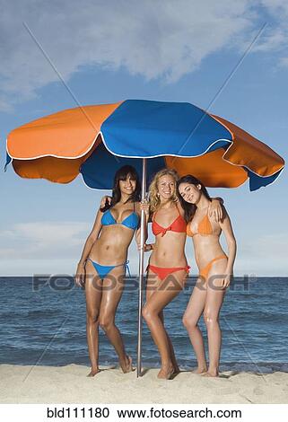 Stock Image - Women in bikinis under beach umbrella. Fotosearch
