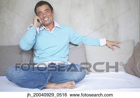 Mid adult man sitting on a bed talking on a mobile phone View Large Photo Image Stock Photograph - Mid adult man sitting on a bed talking on a mobile phone. Fotosearch