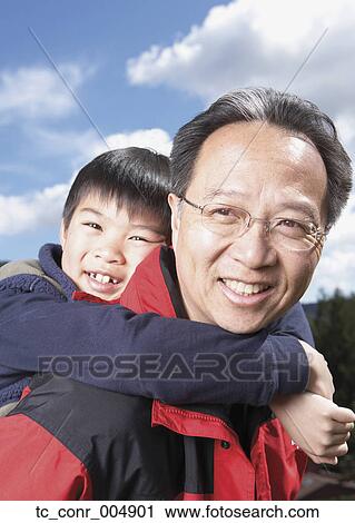 Stock Image - Asian father giving son piggyback ride outdoors. Fotosearch
