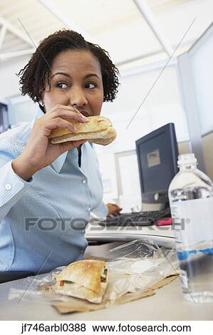 Stock Photo - African American businesswoman eating lunch at desk. Fotosearch