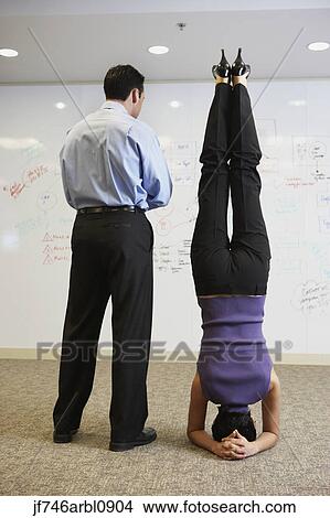 Picture - Businesswoman doing headstand next to businessman in front of whiteboard wall. Fotosearch