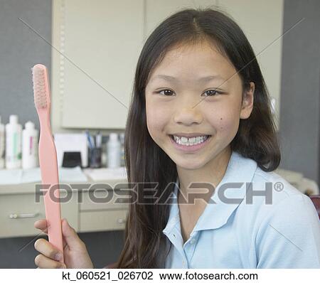 Stock Image - Asian girl holding large toothbrush at dentist's office. Fotosearch