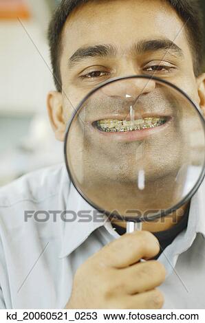 Stock Image - Indian man holding up magnifying glass to his teeth with braces. Fotosearch
