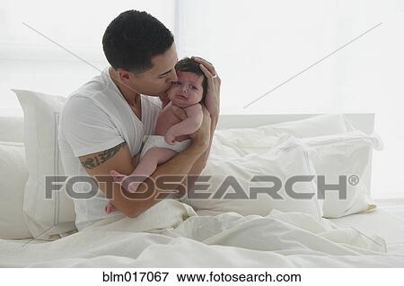 Stock Photo - Hispanic father holding baby in bed. Fotosearch