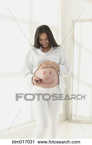 Stock Image - Pregnant Hispanic woman drawing face on her belly. Fotosearch