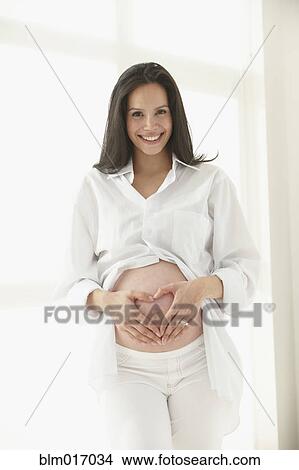 Picture - Pregnant Hispanic woman making heart shape on her belly. Fotosearch