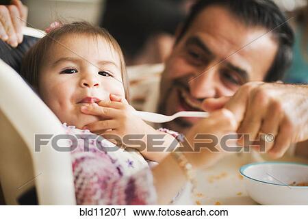 Stock Image - Hispanic father feeding daughter in high chair. Fotosearch
