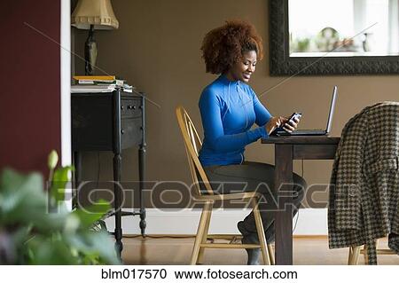 Stock Image - Black woman using cell phone at desk. Fotosearch