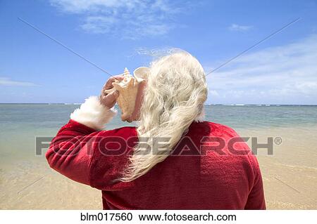 Stock Image - Caucasian Santa Claus listening to seashell on beach. Fotosearch