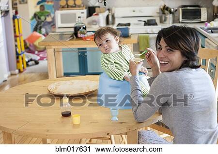 Stock Image - Hispanic mother feeding toddler in kitchen. Fotosearch