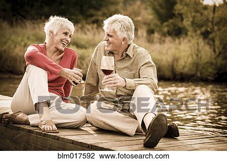 Stock Image - Older Caucasian couple having wine on wooden dock. Fotosearch