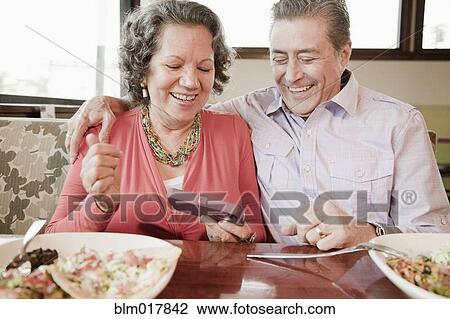 Stock Image - Older Hispanic couple using cell phone in restaurant. Fotosearch