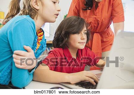 Stock Image - Teacher and children using laptop in library. Fotosearch