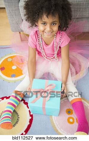 Stock Photo - African American girl opening present in living room. Fotosearch
