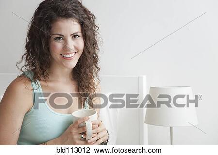 Stock Image - Caucasian woman sitting on bed drinking coffee. Fotosearch