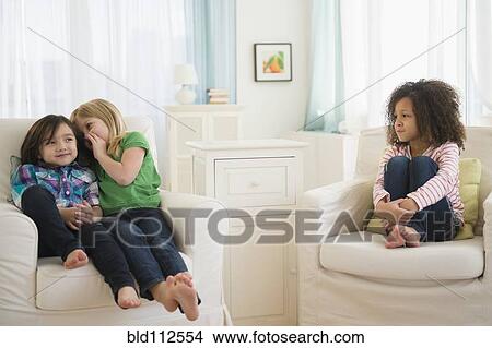 Picture - Girls whispering away from friend in living room. Fotosearch