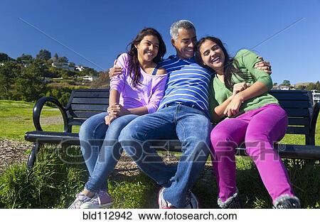 Stock Image - Hispanic father and daughters sitting on park bench. Fotosearch