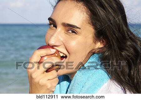 Stock Image - Hispanic woman eating apple near ocean. Fotosearch