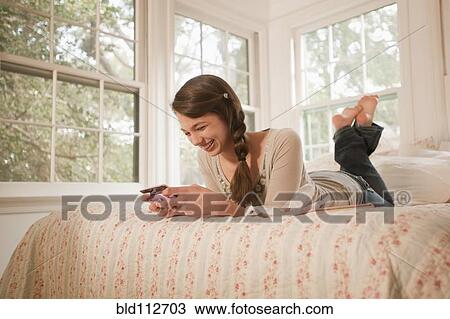 Stock Image - Mixed race girl laying in bed text messaging on cell phone. Fotosearch