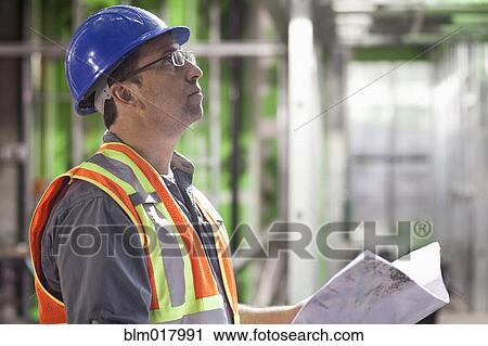 Stock Image - Caucasian worker reading blueprints on site. Fotosearch