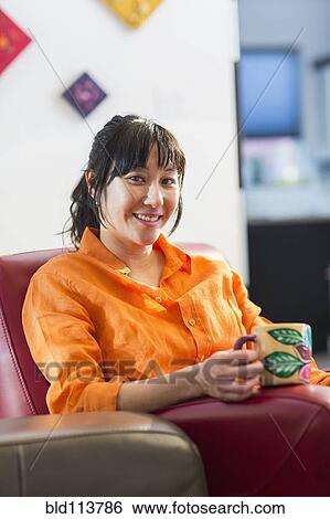 Stock Photograph - Asian woman having cup of coffee in armchair. Fotosearch
