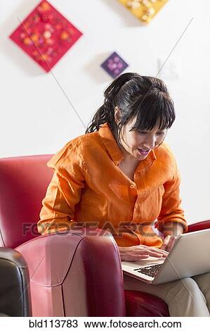 Stock Image - Asian woman using laptop in armchair. Fotosearch
