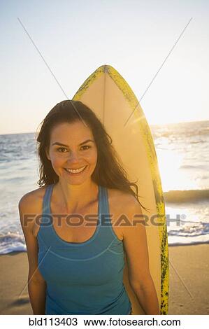 Stock Image - Mixed race woman carrying surfboard on beach. Fotosearch