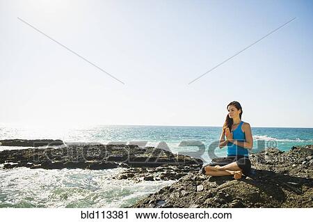 Stock Image - Mixed race woman meditating on rocky beach. Fotosearch