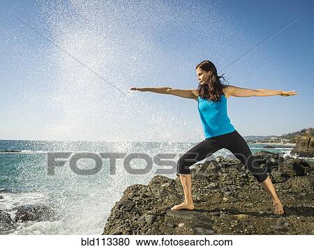 Stock Image - Mixed race woman practicing yoga on rocky beach. Fotosearch
