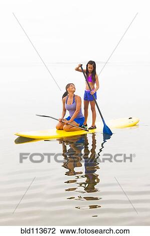 Stock Image - Mother and daughter riding paddle board. Fotosearch